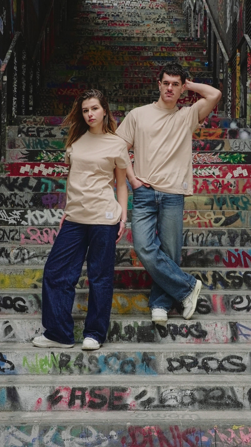 Mannequins homme et femme, portant le t-shirt beige Indieviduals, avec comme décor un escalier rempli de tags. 