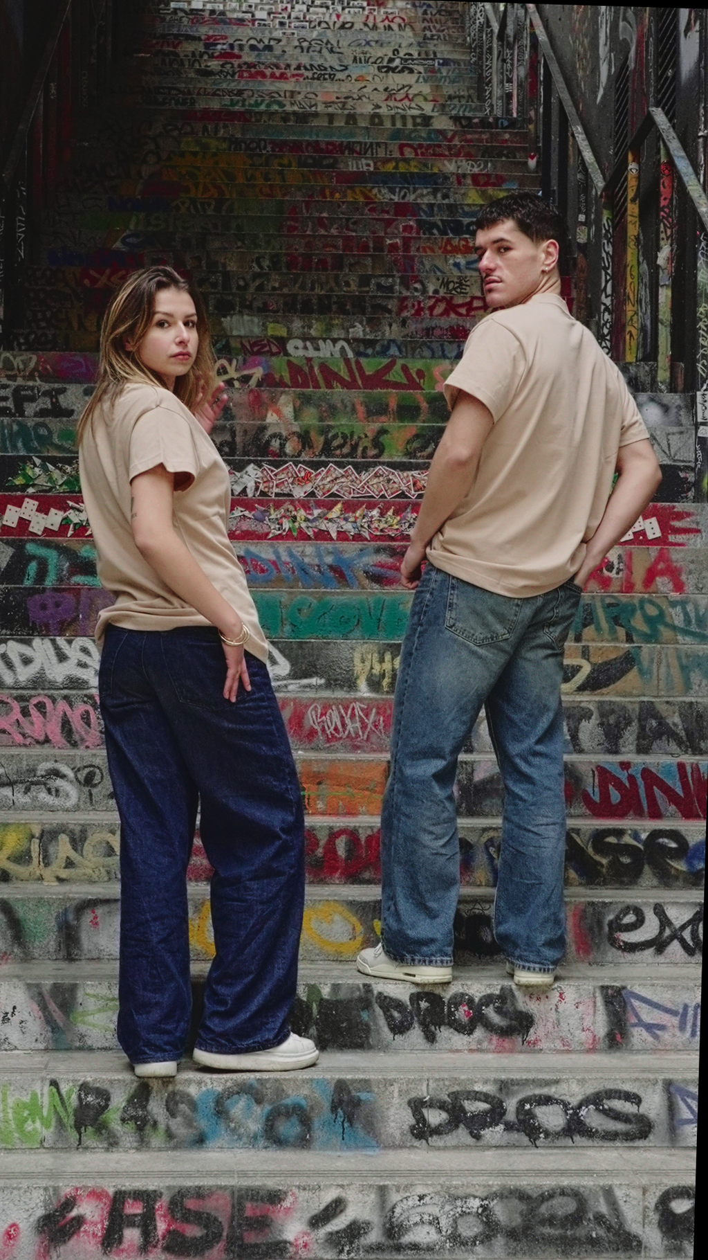 Mannequins homme et femme de dos, portant le t-shirt beige Indieviduals, avec comme décor un escalier rempli de tags. 